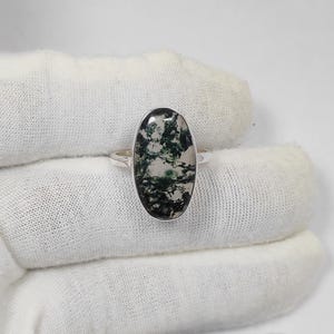 May include: A silver ring featuring an oval-shaped moss agate stone. The stone displays a pattern of green and white inclusions. The ring's band is a simple, polished silver. The ring is set against a white background.