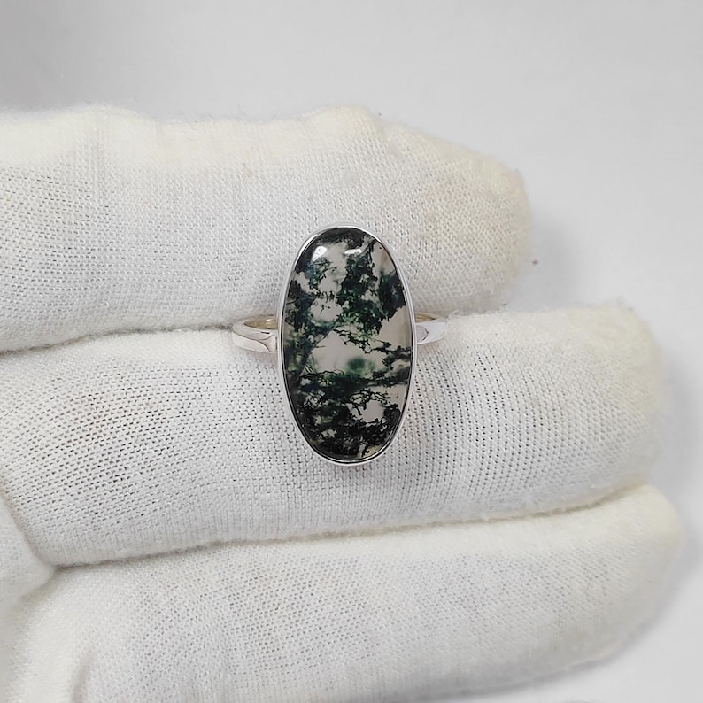 May include: A silver ring featuring an oval-shaped moss agate stone. The stone displays a pattern of green and white inclusions. The ring's band is a simple, polished silver. The ring is set against a white background.