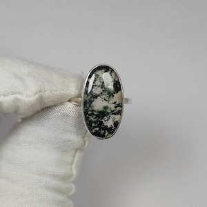 May include: A silver ring featuring an oval moss agate stone. The stone has a translucent base with green and white inclusions, set in a polished silver band. The ring is displayed against a white background.