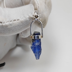 Puede incluir: Un colgante de plata con una piedra preciosa azul. La piedra preciosa es de forma rugosa, natural y está engastada en un engaste de plata.
