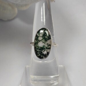 May include: A silver ring featuring an oval-shaped moss agate stone. The stone displays a pattern of green and white, set in a polished silver band. The ring is displayed on a clear stand.