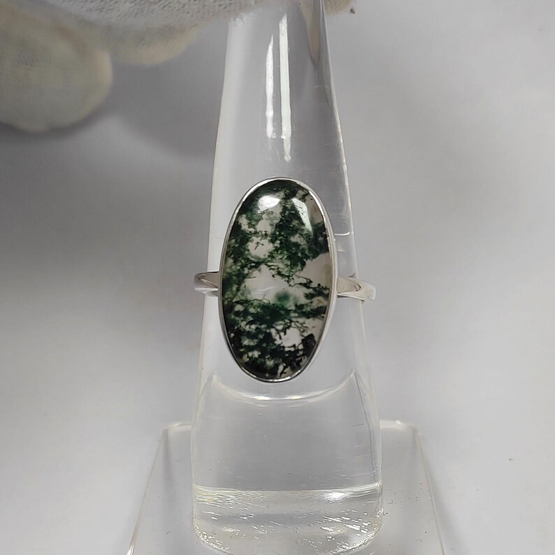 May include: A silver ring featuring an oval-shaped moss agate stone. The stone displays a pattern of green and white, set in a polished silver band. The ring is displayed on a clear stand.