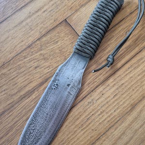May include: A large, gray, metal knife with a paracord handle. The knife has a textured blade and a simple, rounded tip.