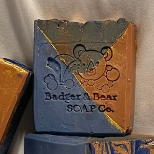 Puede incluir: Tres barras de jabón hechas a mano con un diseño marmoleado azul, marrón y dorado. El jabón está etiquetado como "Badger & Bear Soap Co."