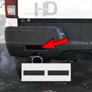 May include: A white pickup truck with a black rear bumper. The bumper has a red reflector on the left side. The truck is parked on a black asphalt surface.