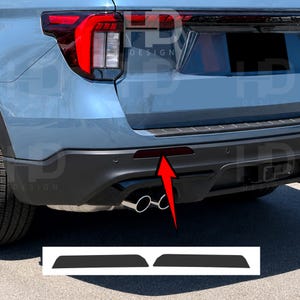 May include: A light blue vehicle rear bumper featuring a red reflector highlighted by a red arrow. Below the vehicle, two dark, trapezoidal adhesive overlays are displayed on a white background, designed to cover the rear bumper reflector area.