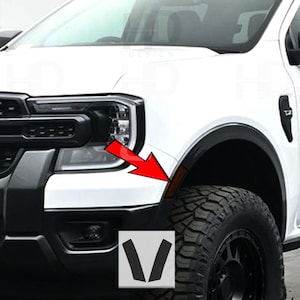 May include: White truck with black accents, a large tire, and a black fender flare. The image includes a red arrow pointing to a small amber-colored light. A white square with black V-shaped graphics is in the lower corner.