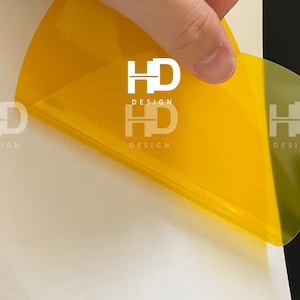 May include: A yellow, semi-transparent sticker with the text "HD DESIGN" printed in white. The sticker is being peeled off of a white surface.