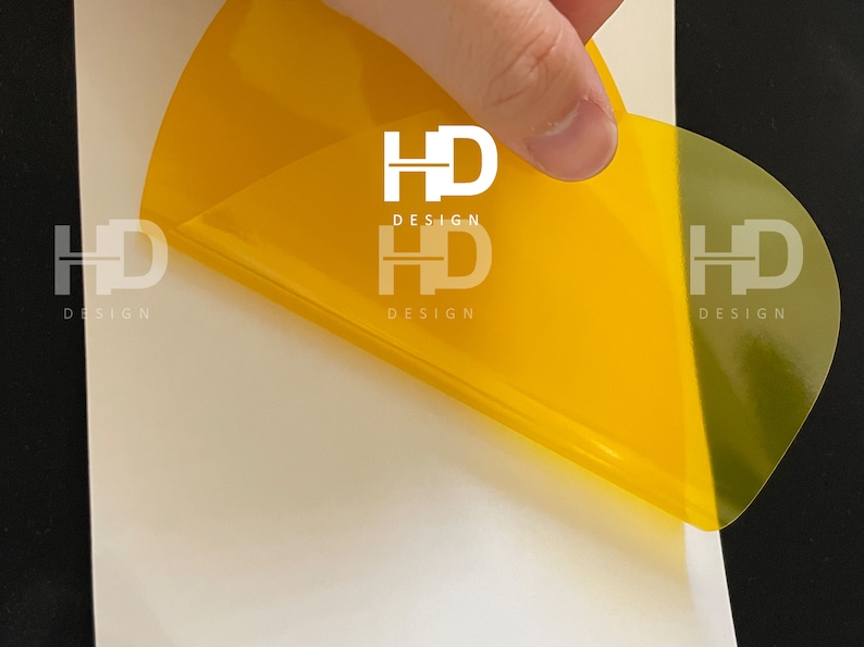 May include: A yellow, semi-transparent sticker with the text "HD DESIGN" printed in white. The sticker is being peeled off of a white surface.
