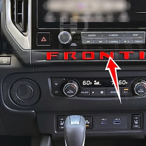May include: Close-up of a car dashboard with the word "FRONTIER" in red letters. The dashboard is black with various buttons and controls, including climate control and a display screen. The temperature is set to 60 degrees Fahrenheit.