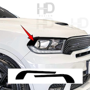 May include: A white vehicle front end featuring a black headlight eyelid decal applied to the upper edge of the headlight assembly. A red arrow points to the decal, which is also shown as a separate black graphic element at the bottom of the image.