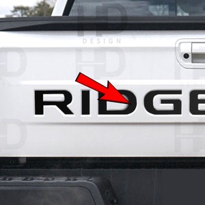 May include: A white pickup truck with a black "RIDGE" tailgate decal. The decal is centered on the tailgate and is in all capital letters.