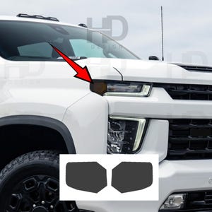 May include: A white pickup truck front end featuring a red arrow pointing to the amber side marker light. Below the truck, two dark gray, rectangular adhesive overlays are displayed, designed to cover the side marker lights on the vehicle.