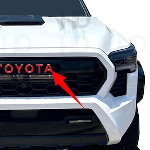 May include: The front end of a white Toyota truck featuring a black honeycomb grille with the word TOYOTA in red lettering. A prominent red arrow points toward the letter A in the brand name. The vehicle includes dark headlights and a hood scoop.