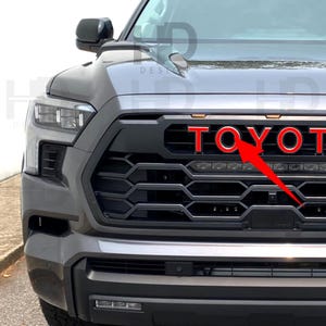May include: Close-up of a gray Toyota truck front end. The black grill features the red word "TOYOTA". The truck has black accents and a black bumper. The headlights are visible.