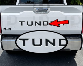 May include: A white pickup truck with a black "TUNDRA" emblem on the tailgate. The emblem is raised and has a 3D effect.