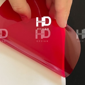 May include: A red vinyl sticker with the text "HD DESIGN" in white letters. The sticker is being peeled off of a white surface and is partially on a black surface.