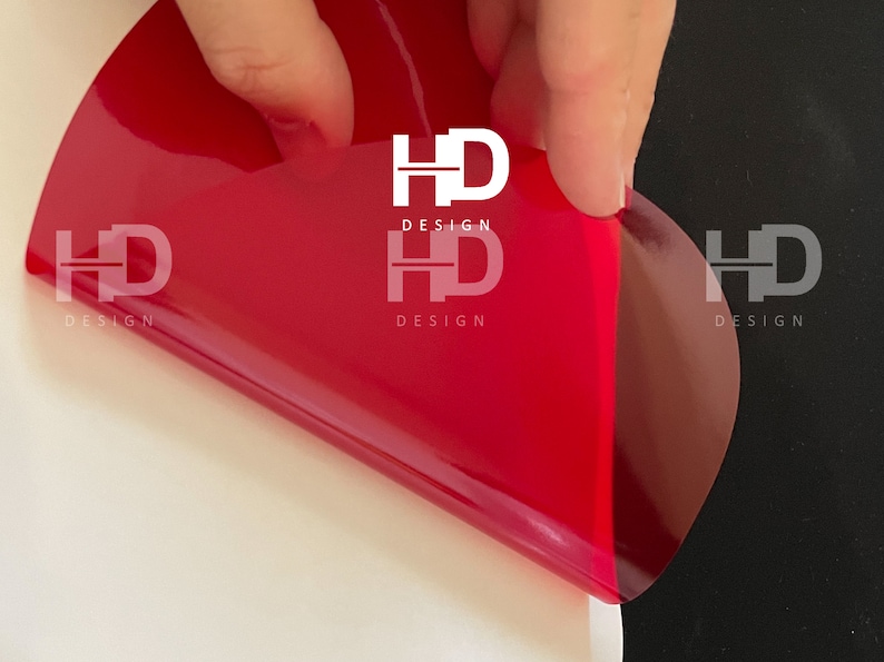 May include: A red vinyl sticker with the text "HD DESIGN" in white letters. The sticker is being peeled off of a white surface and is partially on a black surface.