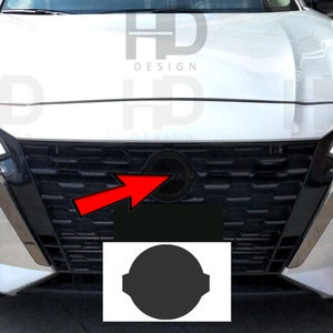 May include: A silver car with a black grille and a Nissan logo. The grille has a honeycomb pattern and the logo is centered in the grille. The car is parked in a parking lot.