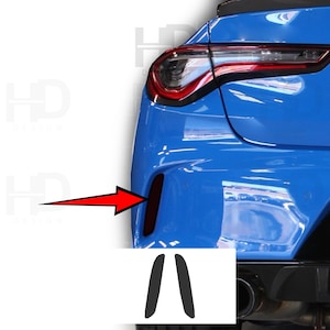 May include: A close-up of a blue car's rear bumper, with a black accent and a red arrow pointing to a dark vent. Below, two black, curved pieces are displayed, likely car accessories.