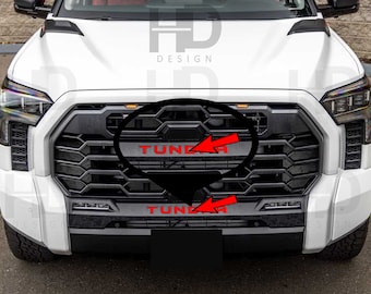 May include: Front view of a white truck with a black grille. The word "TUNDRA" is in red on the grille. The design includes a black lower bumper and headlights.