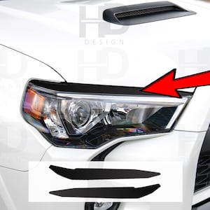 May include: A set of two black vinyl overlays for a white car. The overlays are designed to cover the lower portion of the headlights and add a sporty look to the vehicle. The overlays are shown on a white car with a black grille and black trim.