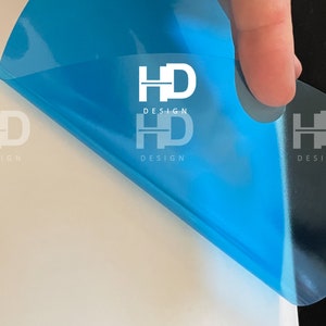 May include: A blue transparent sticker with the text "HD DESIGN" printed in white. The sticker is being peeled off of a white surface.