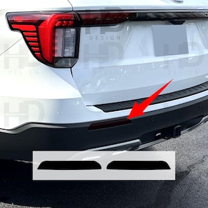 May include: Black bumper reflector overlays for a white SUV. The image shows the rear of the vehicle with the taillight and bumper details. Two black overlays are shown below the bumper, ready for installation. The word "DESIGN" is visible.