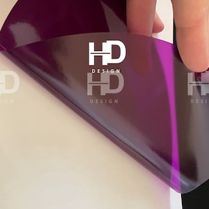 May include: A close-up of a purple vinyl sticker with the text "HD DESIGN" printed on it. The sticker is being peeled off of a white surface.