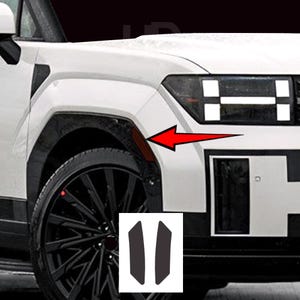 May include: Close-up of a white vehicle's front quarter panel, featuring a black wheel and tire. A red arrow points to a dark red accent piece. Two dark gray, rectangular decals are in the lower right corner.
