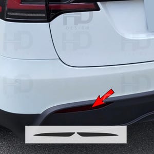 May include: A close-up of the rear bumper of a white Tesla Model Y with black trim. The rear bumper has a red reflector and two black rectangular cutouts for rear reflectors. The text "HD DESIGN" is visible on the rear bumper.