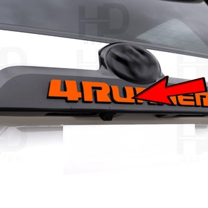 May include: A black rear hatch trim with an orange "4RUNNER" emblem. The emblem is on the left side of the trim, and there is a black camera lens on the right side.