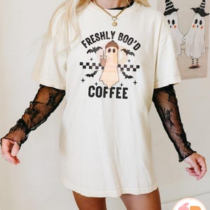 May include: Cream-colored t-shirt with the text "FRESHLY BOO'D COFFEE" and a ghost graphic holding a coffee cup. The design includes bats and a checkered pattern. The shirt is worn with a black lace long sleeve shirt.