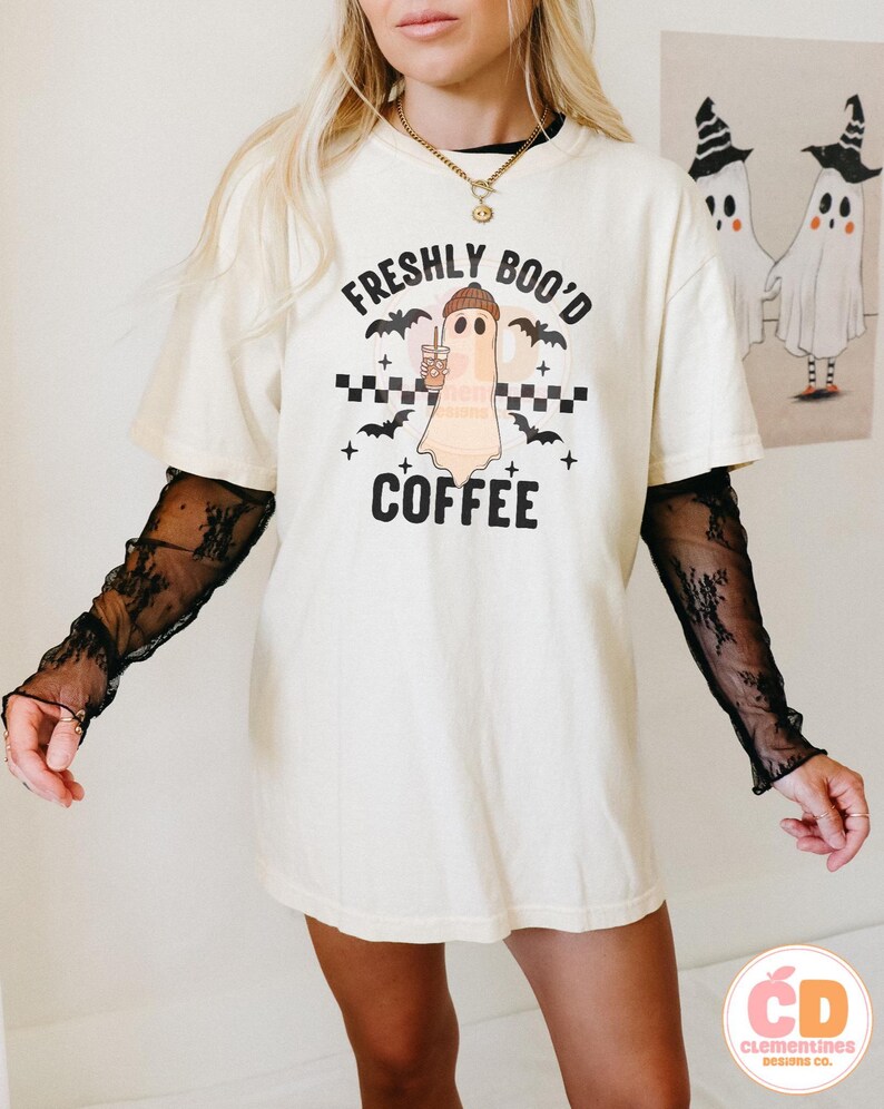 May include: Cream-colored t-shirt with the text "FRESHLY BOO'D COFFEE" and a ghost graphic holding a coffee cup. The design includes bats and a checkered pattern. The shirt is worn with a black lace long sleeve shirt.