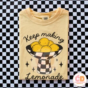 May include: Yellow t-shirt with a graphic of lemons in a black and white checkered bowl. The text "Keep making Lemonade" is above and below the bowl. The shirt has a crew neck and short sleeves.