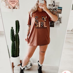 Puede incluir: Una camiseta de color &oacute;xido con el texto "COW GIRL" en una mezcla de fuentes y patrones. La modelo lleva botas de vaquero en blanco y negro. Un cactus en una maceta blanca es visible en el fondo.