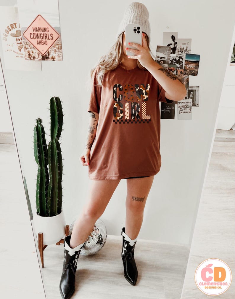 Puede incluir: Una camiseta de color &oacute;xido con el texto "COW GIRL" en una mezcla de fuentes y patrones. La modelo lleva botas de vaquero en blanco y negro. Un cactus en una maceta blanca es visible en el fondo.