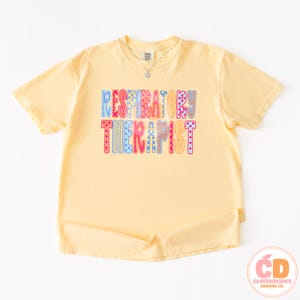 May include: A light yellow t-shirt with the words "RESPIRATORY THERAPIST" in colorful, patterned letters. The text is in a playful font with blue, pink, and yellow accents. A small pendant necklace is visible.