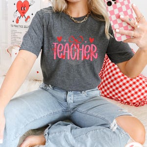 May include: A dark gray t-shirt with the text "XO TEACHER" in pink with hearts and glitter. The model is wearing blue jeans and holding a pink and white checkered phone case.