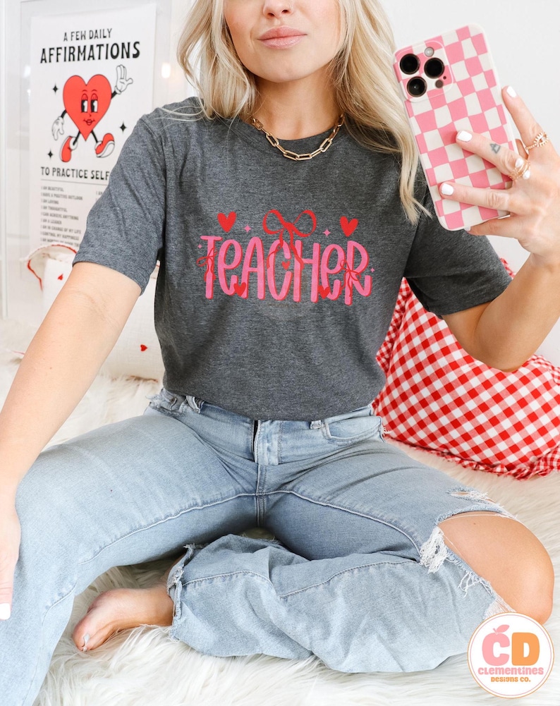 May include: A dark gray t-shirt with the text "XO TEACHER" in pink with hearts and glitter. The model is wearing blue jeans and holding a pink and white checkered phone case.