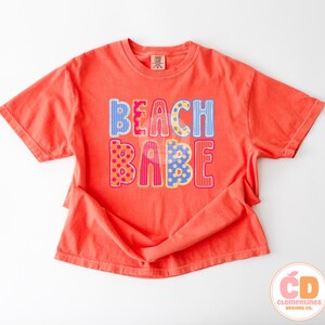 May include: Coral-colored t-shirt with the words "BEACH BABE" in colorful, patterned letters. The letters are decorated with stripes, polka dots, and other designs. The shirt has short sleeves and a crew neck.