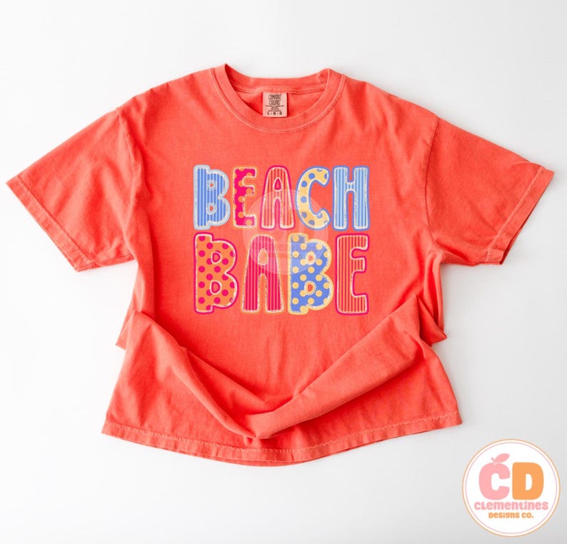 May include: Coral-colored t-shirt with the words "BEACH BABE" in colorful, patterned letters. The letters are decorated with stripes, polka dots, and other designs. The shirt has short sleeves and a crew neck.