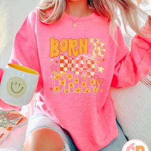 May include: A bright pink sweatshirt with the text "BORN TO DILLY DALLY" in orange and yellow. The design includes stars and a checkered pattern. A white mug with a yellow interior and a smiley face is held in the image.