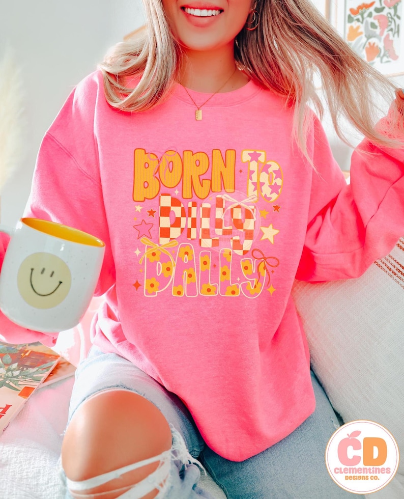 May include: A bright pink sweatshirt with the text "BORN TO DILLY DALLY" in orange and yellow. The design includes stars and a checkered pattern. A white mug with a yellow interior and a smiley face is held in the image.