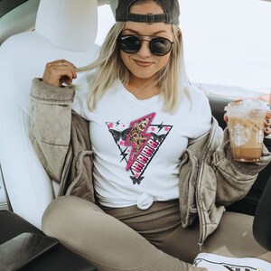 Pu&ograve; includere: Una donna indossa una maglietta bianca con un fulmine rosa e la parola "Warrior" in lettere nere. Indossa anche un cappello da baseball nero, occhiali da sole, una giacca marrone e pantaloni marroni.