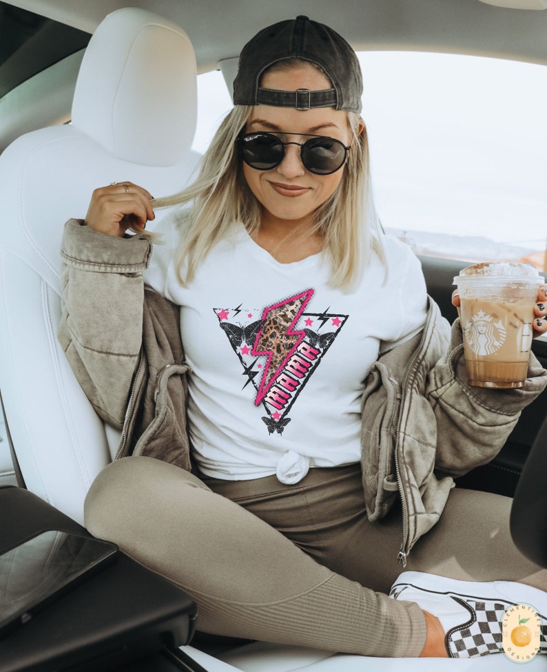 Pu&ograve; includere: Una donna indossa una maglietta bianca con un fulmine rosa e la parola "Warrior" in lettere nere. Indossa anche un cappello da baseball nero, occhiali da sole, una giacca marrone e pantaloni marroni.