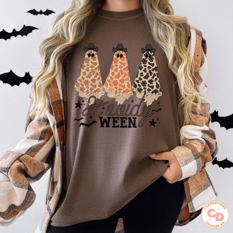May include: Brown t-shirt with a Halloween-themed graphic. The design features three ghost figures wearing cowboy hats, each with a different pattern: cow print, orange, and black and white. The text "Howdy Ween" is below the ghosts.