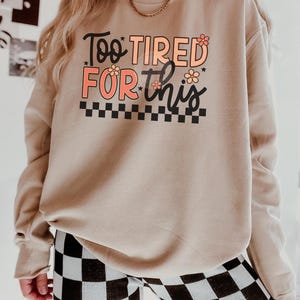May include: A beige sweatshirt with the text "Too Tired For This" in black and pink lettering with small flower accents. The text is surrounded by a black and white checkered pattern.