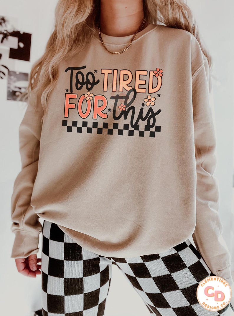 May include: A beige sweatshirt with the text "Too Tired For This" in black and pink lettering with small flower accents. The text is surrounded by a black and white checkered pattern.