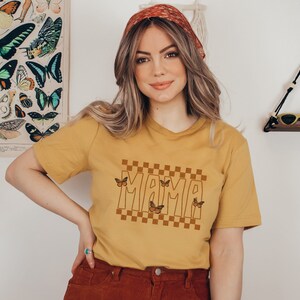 Puede incluir: Una camiseta amarilla mostaza con un patr&oacute;n a cuadros y la palabra "MAMA" en una fuente retro. Hay varios gr&aacute;ficos de mariposas marrones en la camiseta.